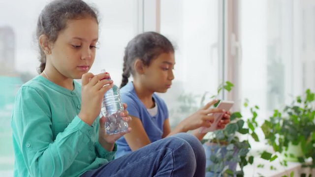 Handheld shot of cute little girl sitting on window sill and drinking water as he friend browsing internet on tablet during recess in primary school