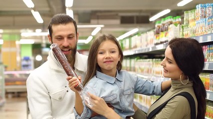 Young happy family are buying groceries in the supermarket. Mom dad and daughter buy sausage in the store, dad holds her daughter in his arms and they pretend that they bite the sausage. Slow motion.