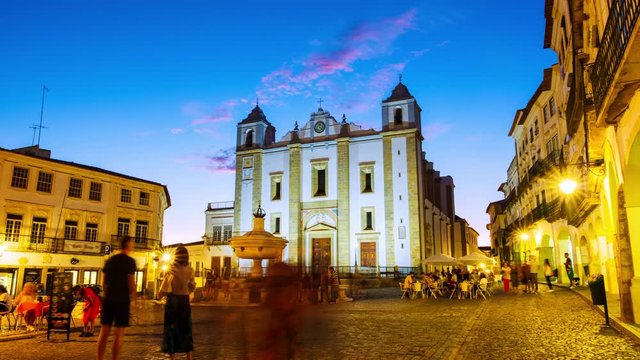 Evora, Portugal. View of Roman Catholic Cathedral of Evora, Portugal at sunset. Illuminated historical buildings at the square with motion blurred people. Time-lapse of sky gettind dark