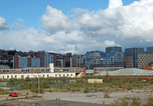 a large empty brownfield site in leeds england with weeds growing through concrete and a burned out abandoned car surrounded by city apartment buildings