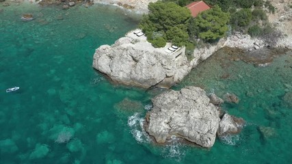 Flying over a cape on which is seen a lonely house surrounded by coniferous trees. The turquoise sea washes the rocky coast of the cape and the sandy coastline, sea waves roll on the shore