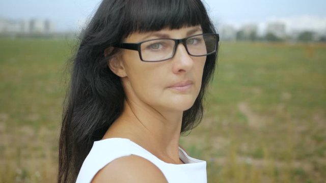 Young woman with in white dress on a windy day on the outskirts of the city of New Yor.  Portrait of a businesswoman with glasses looking at the camera. Closeup. Slow Motion.