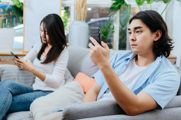 asian young couple man and woman absorbed and focused into mobile phone and sitting on sofa in living room at home, social media addiction, upset couple, family issues and relationship concept
