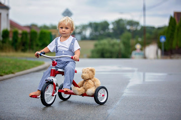 Cute little boy, with teddy bear toy, riding tricycle on the street in the rain, barefeet