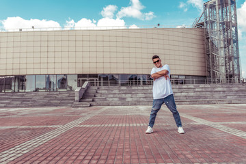 sporty male dancer, standing in pose of confidence, young guy, free space for text of moticavia, in summer in city against the background of steps and a building, jeans sunglasses, white T-shirt