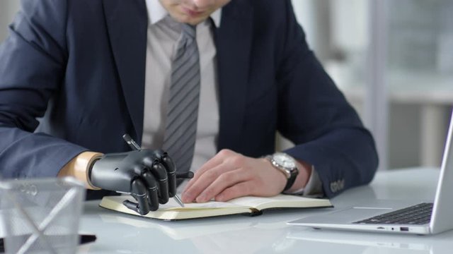 Close up shot of businessman with prosthetic bionic hand writing with pen in notepad at office desk