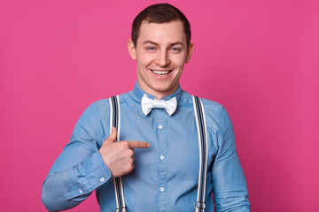 Portrait of stylish handsome young man posing isolated on rosy background. Handsome male smiling and pointing aside with his index finger, guy wearing blue shirt, suspenders and white bow tie.