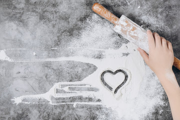Baking background. Girl hands keep rolling pin and heart of flour on gray table with copy space, top view. Valentine's Day