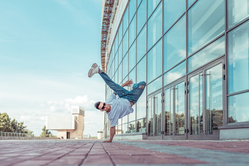strong sporty man dancing break dance, standing one arm, young guy, free space for text motivation lifestyle, summer city against background glass building window, jeans, sunglasses, a white T-shirt
