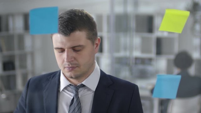 Caucasian businessman with prosthetic hand attaching sticke notes to glass wall in the office and reading tasks on them