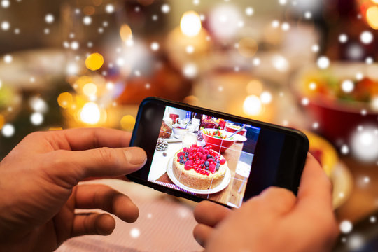 food, technology and holidays concept - close up of male hands photographing cake by smartphone at christmas dinner over snow