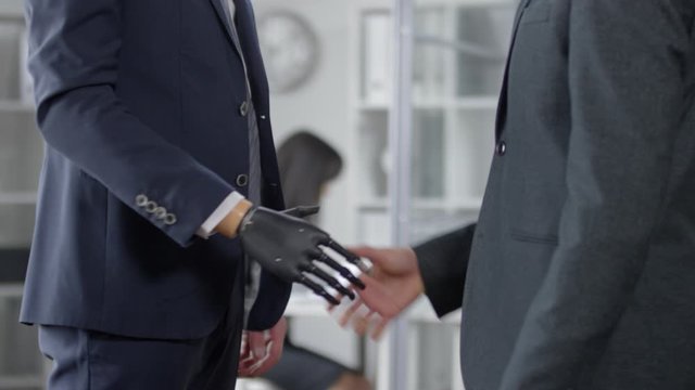 Mid-section shot of businessman with prosthetic hand standing in the office, giving handshake to male colleague and discussing something with him