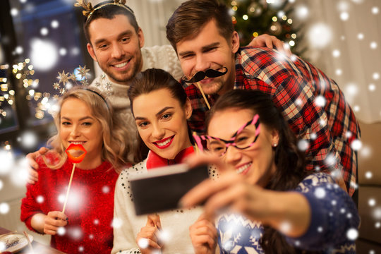 holidays and celebration concept - happy friends with party props taking selfie by smartphone at home christmas dinner over snow