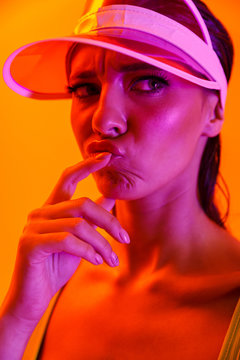 Image closeup of fashionable brunette woman wearing sun visor cap and sportswear posing at camera isolated over orange neon wall