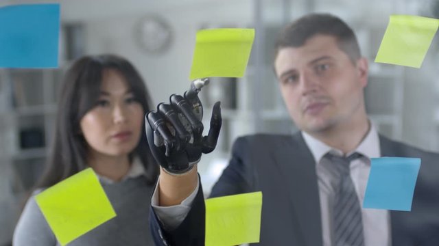 View through glass wall of caucasian businessman with prosthetic hand explaining tasks on sticky notes to Asian female colleague while discussing work in office