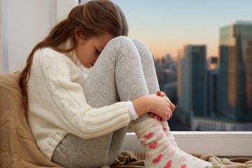 childhood, sadness and people concept - sad beautiful girl in sweater sitting on sill at home...