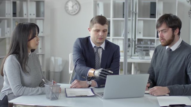 Caucasian businessman with prosthetic hand sitting office table, pointing on laptop screen and explaining contract to multiethnic colleagues during corporate meeting