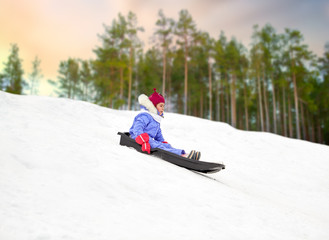 childhood, sledging and season concept - happy little girl sliding on sled down snow hill outdoors...