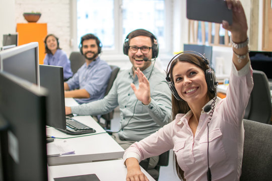 Beautiful woman customer support operator taking a selfie photo of her team colleagues in a call center.