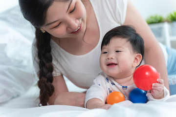 Young beautiful asian mother with asian baby on bed and playing toy ball together on white bed with feeling happy and cheerful and the baby that crawling on the bed.Baby family concept