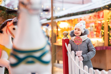 Fototapeta premium holidays, childhood and people concept - happy little girl with big lollipop at christmas market