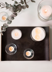 decoration, hygge and cosiness concept - burning white fragrance candles on tray and branches of eucalyptus populus on table