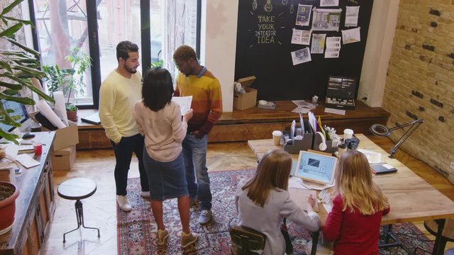 High angle view of two female colleagues discussing interior plan on digital tablet at table while diverse colleagues discussing papers in modern loft office