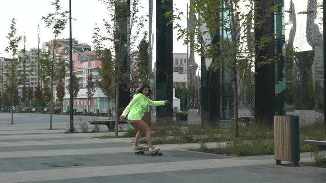  A beautiful girl from Russia is riding a skateboard along the street. Modern girl in motion. She is wearing a bright jacket with a light green hood.