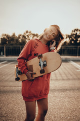 Beauty portrait of active girl with skateboard on road near parking in summer sunset in evening. Stylish urban woman model posing in red hoodie, sneakers and longboard © Yevhen