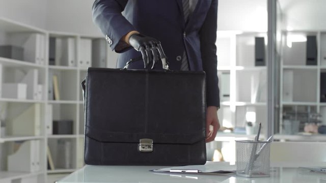 Caucasian businessman with prosthetic bionic hand walking to his workplace, putting briefcase on office desk and then sitting and typing on laptop