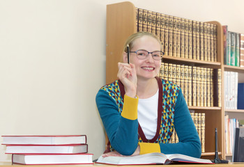 A smiling beautiful woman teacher or librarian at workplace with big books on the background of bookshelves. Copy space