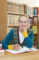 A smiling beautiful woman teacher or librarian at workplace with big books on the background of bookshelves. Copy space