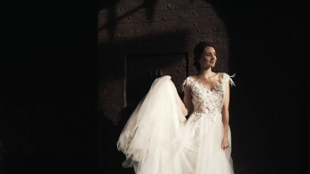 Close-up view of young beautiful bride in a fashionable white wedding dress standing and smiling near old dark wall and gates. Action. A storybook wedding