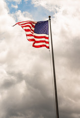 American flag waving atop a flagpole against a cloudy darkening grey sky towards bottom of frame and a patch of clear blue sky just above "Old Glory".