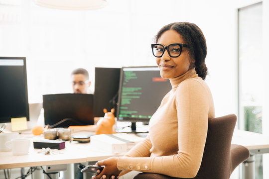 Portrait of female programmer sitting by desk with colleague in background at creative office