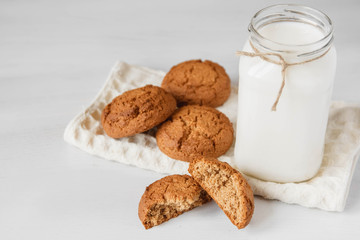 Milk in glass jar and oatmeal cookies near napkin on white table background. Copy, empty space for text