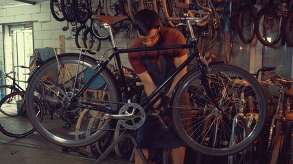 theme small business bike repair. young Caucasian brunette man wearing safety goggles, gloves and apron uses hand tool repairing and adjusting crank and pedal system bicycle in the garage workshop