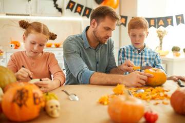 Young father sitting at the table together with his two children and they preparing pumpkins for Halloween party at home