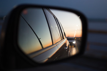 side mirror of a car in which a road with a traveling car is reflected at sunset