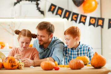 Fototapeta premium Cute girl and boy decorating ripe pumpkins together with their father while sitting at the table at home