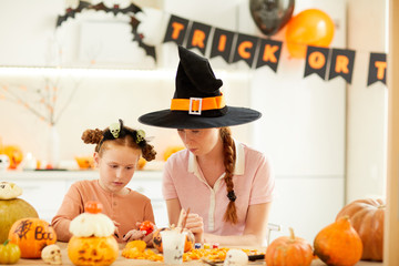 Young mother in witch's hat sitting at the table among pumpkins together with her daughter and they making crafts at home