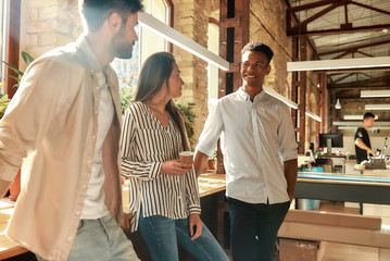 Pause at work. Young and cheerful colleagues in casual wear talking about something and holding coffee cups while standing in the modern office. Coffee break