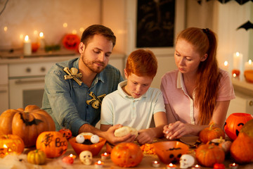 Mother and father sitting at the table together with their son and helping him to make a lantern from pumpkins for Halloween