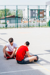 Two strong caucasian athletes resting on the ground at the basketball court while having a conversation.