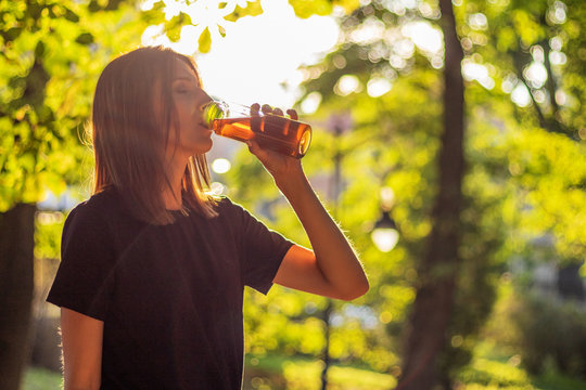 Beautiful fitness athlete woman are drinking iced tea after workout exercising on sunset evening in park. Outdoor portrait with sun glares.