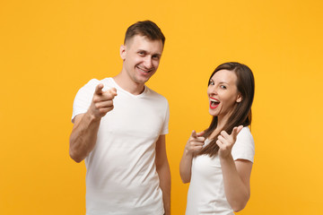 Funny young couple two friends guy girl in white empty blank design t-shirt posing isolated on yellow orange background. People lifestyle concept. Mock up copy space. Pointing index fingers on camera.