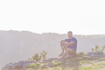 Naklejka na ściany i meble Handsome athlete sitting at a rocky peak while looking at the breathtaking mountain line and a beautiful lake while wearing a blue shirt and grey shorts.