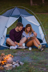 Young couple drinking coffee, enjoying the mountain view and checking the tablet with bonfire in the sunset