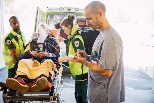 Male nurse writing report while paramedics pushing patient on gurney in hospital