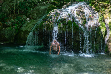A man stands under the streams of water of a mountain waterfall